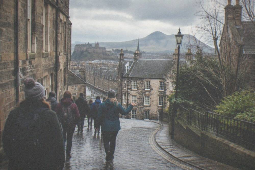 A stone street in Edinburgh curving downhill toward distant hills under an overcast sky