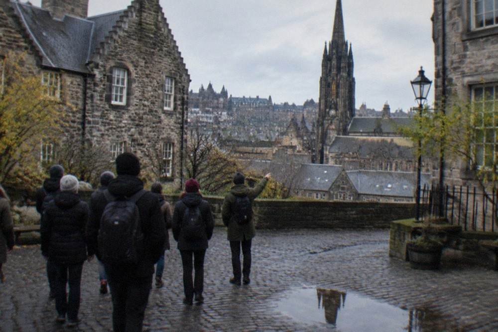 A quiet Edinburgh courtyard of grey stone buildings seen from a cobbled lane, with a church tower beyond the rooftops