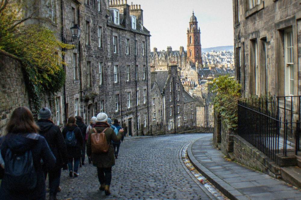 A cobbled Edinburgh street of tall grey tenements curving downhill, with shopfronts and a stone bell tower above the rooftops