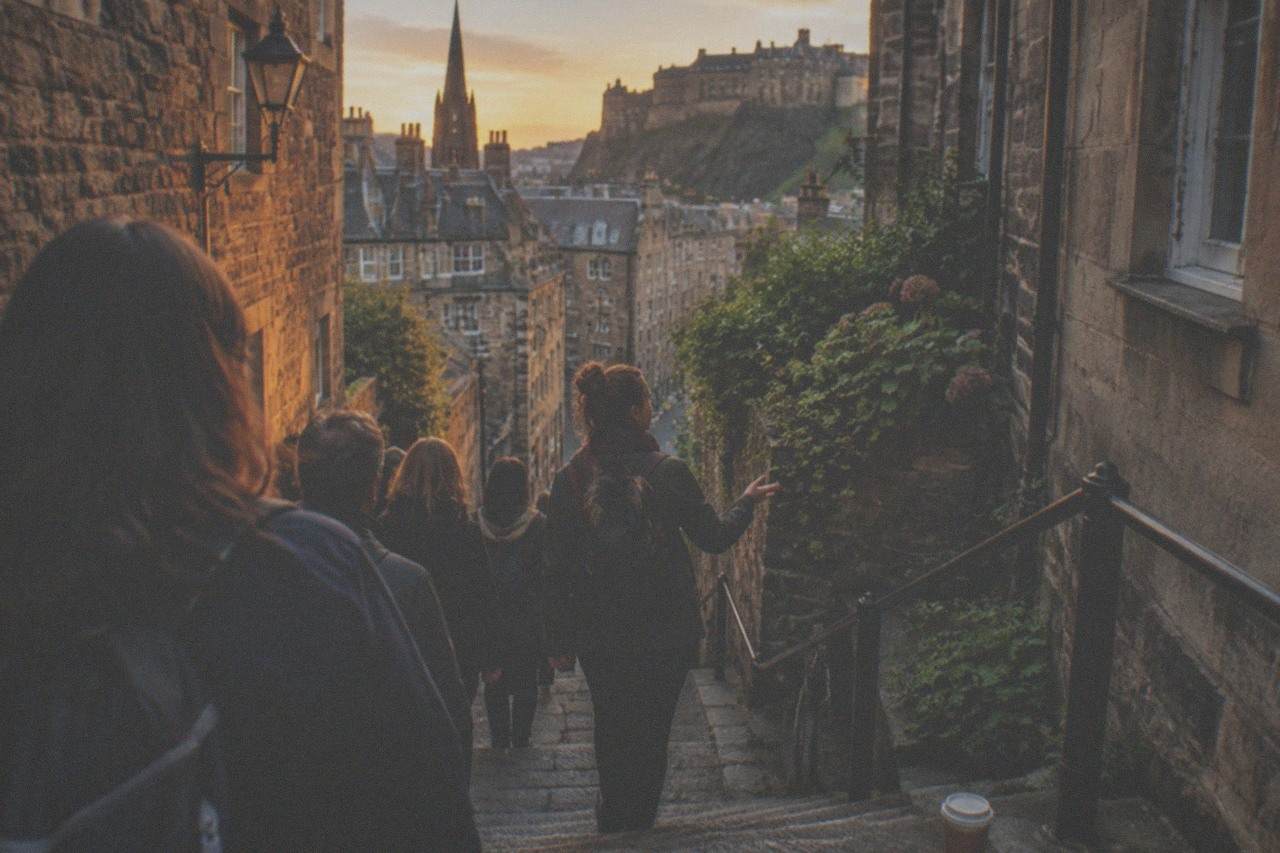 A narrow Edinburgh close between old stone buildings, with worn steps leading downhill toward a sunlit street