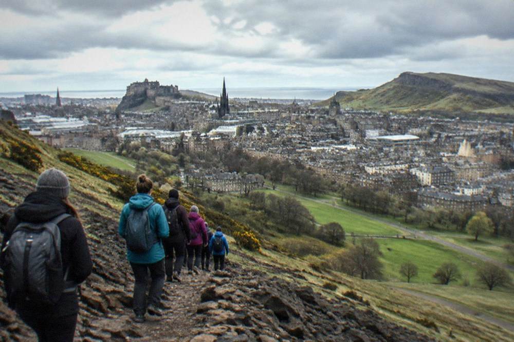 View from Arthur's Seat looking down over Holyrood Park toward the city, with the Firth of Forth in the distance under a cloudy sky.