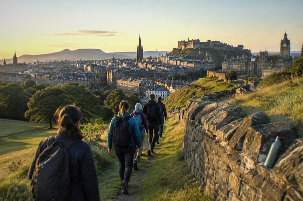 A small group of visitors listening to a guide beside a stone wall on an Edinburgh hillside path
