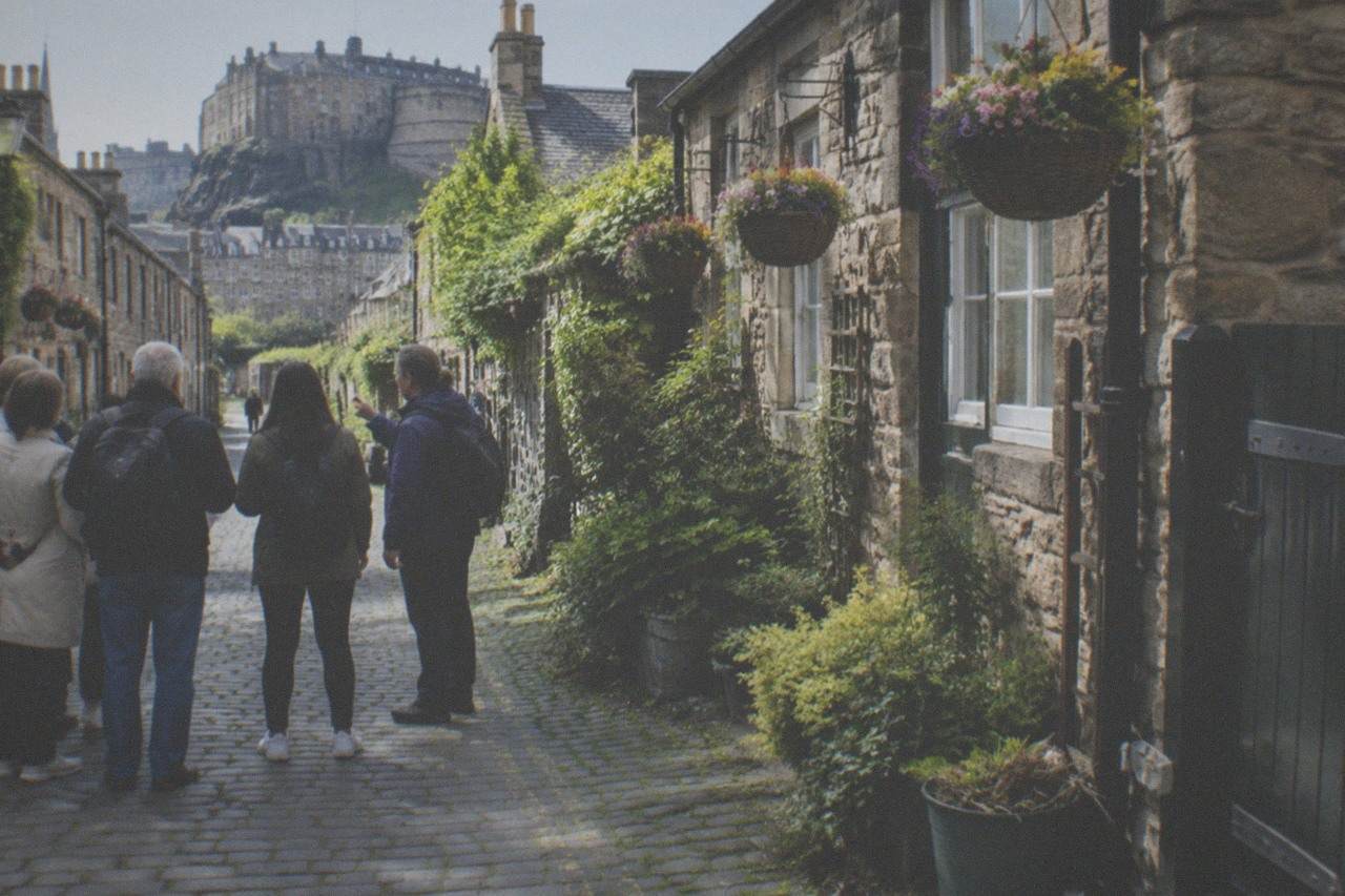 A narrow cobbled lane in Edinburgh lined with small stone houses and hanging baskets beside a quiet stream