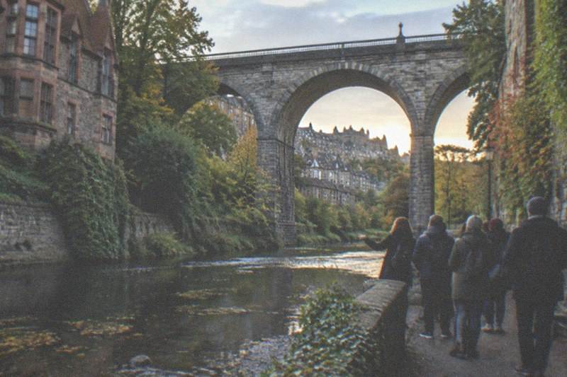 Stone cottages and the stream of the Water of Leith beneath the tall arches of a Dean Village bridge, ivy on the old walls, one walker on the waterside path in evening light.