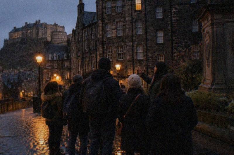 A narrow Old Town close after dark, wet cobbles catching amber lamplight below towering blackened tenements, a single lit window high in the stone wall.
