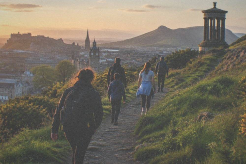 A green hillside path climbing toward a rocky summit above a city