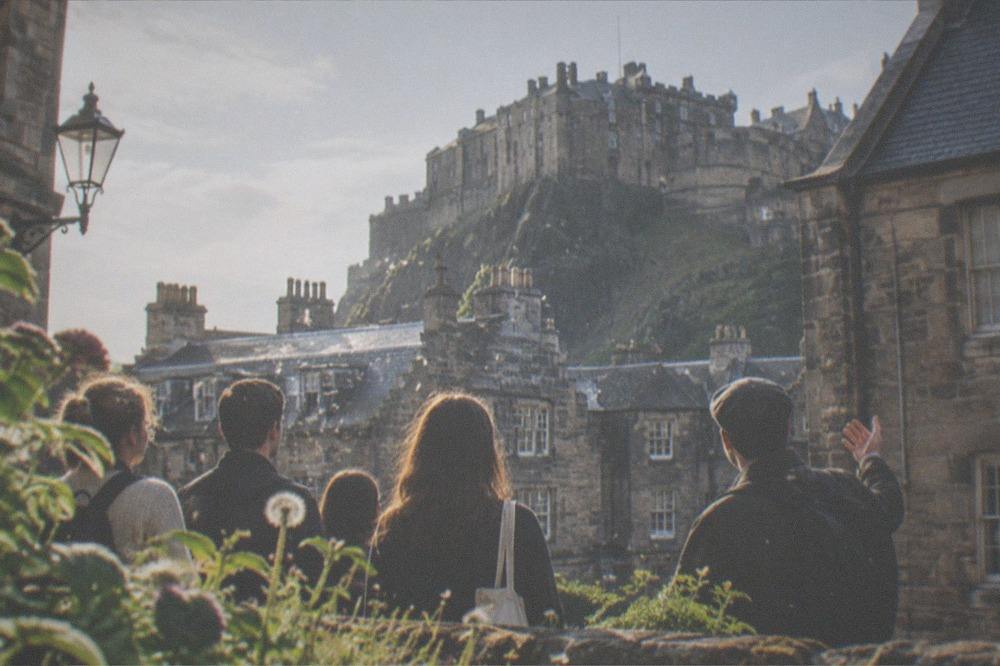 A stone castle on a rocky crag above city rooftops seen from a garden below