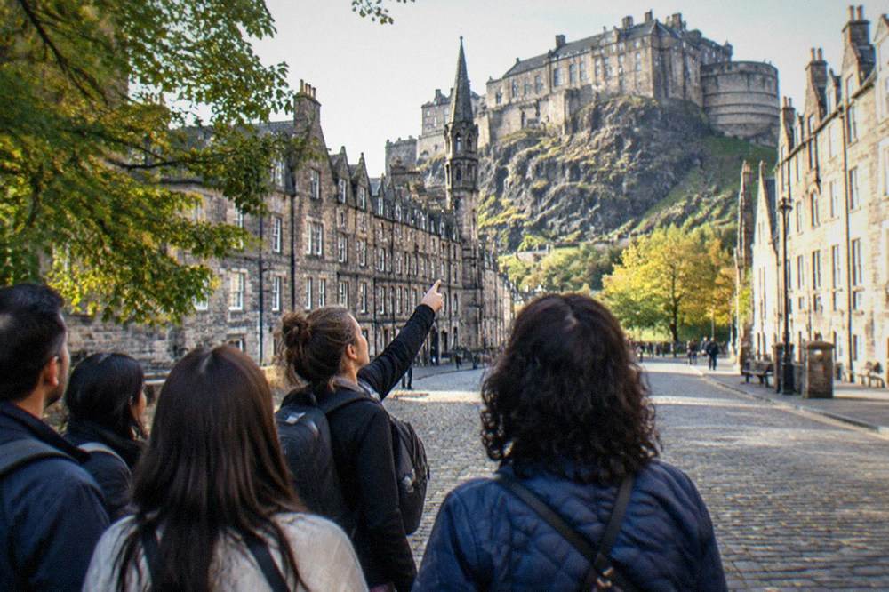 A Spanish-speaking guide gesturing towards Edinburgh Castle from the Royal Mile with a tour group listening