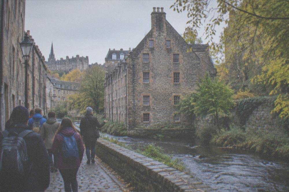 A stone village lane beside a river with a tall gable-ended building and trees